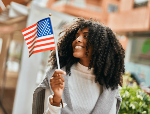 Mulher negra com cabelos longos e encaracolados, vestindo roupas brancas, segura uma pequena bandeira dos Estados Unidos enquanto caminha ao ar livre, sorrindo.